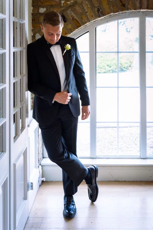 A handsome groom in a sharp black suit and bow tie poses casually against a doorway with natural window light illuminating the scene.