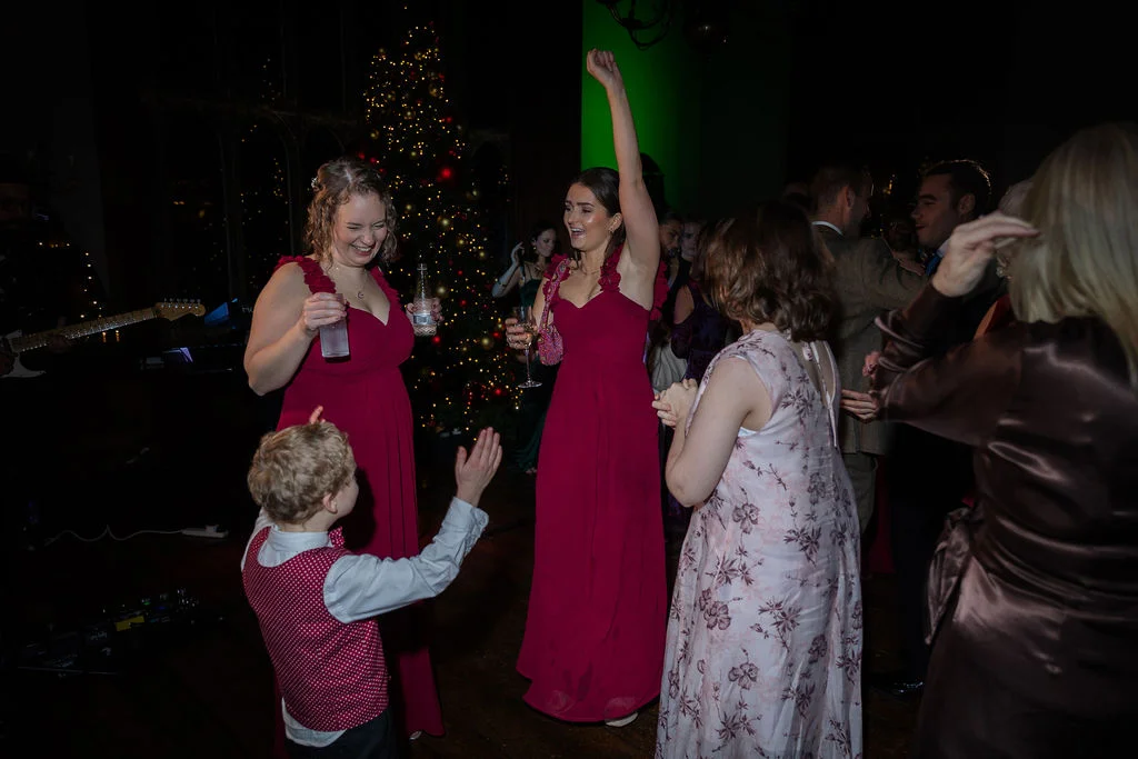 Wedding guests and a young page boy dance enthusiastically on the dance floor in front of a festive Christmas tree during an evening reception.