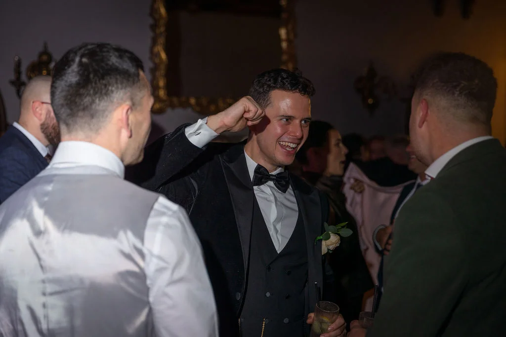 A groom in a black tuxedo and bow tie laughs and raises his fist in celebration while mingling with guests at an evening reception.