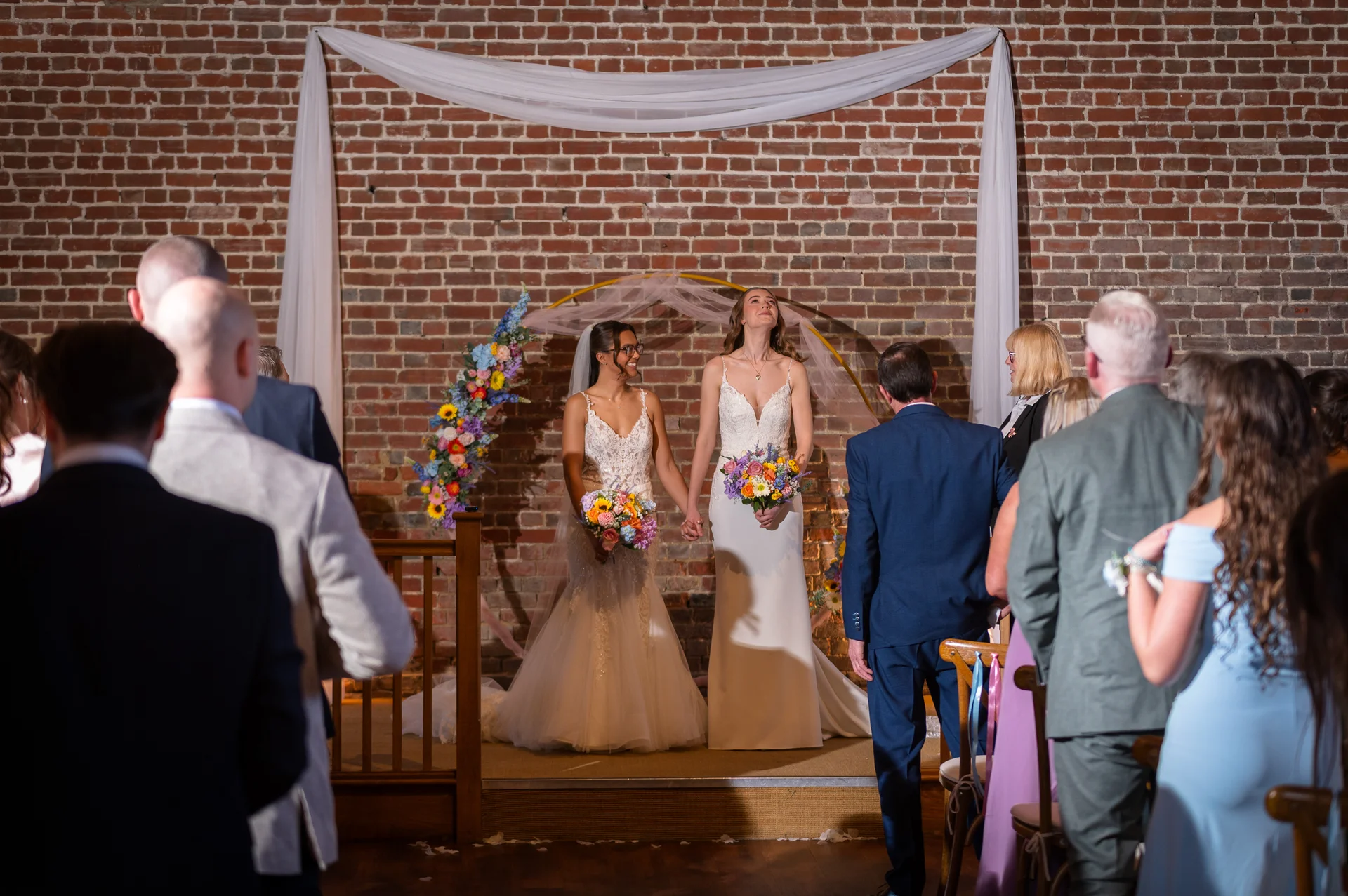 Two brides stand together holding bouquets during their ceremony in front of an exposed brick wall adorned with white drapery and colourful floral arrangements.