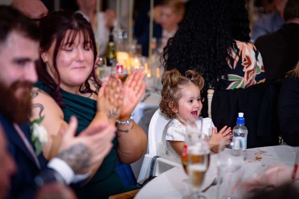 A young toddler sits in a high chair at a wedding reception table, laughing joyfully and clapping along with surrounding guests during speeches.