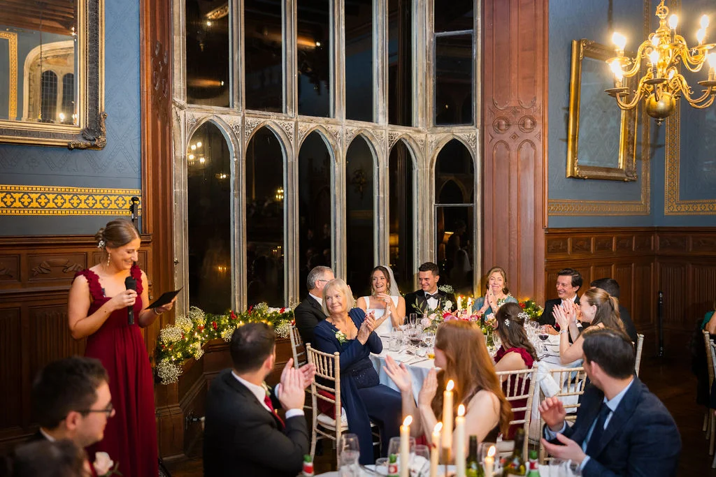 A bridesmaid in a burgundy dress delivers a speech in a grand, wood-panelled hall with tall windows as guests laugh and applaud.