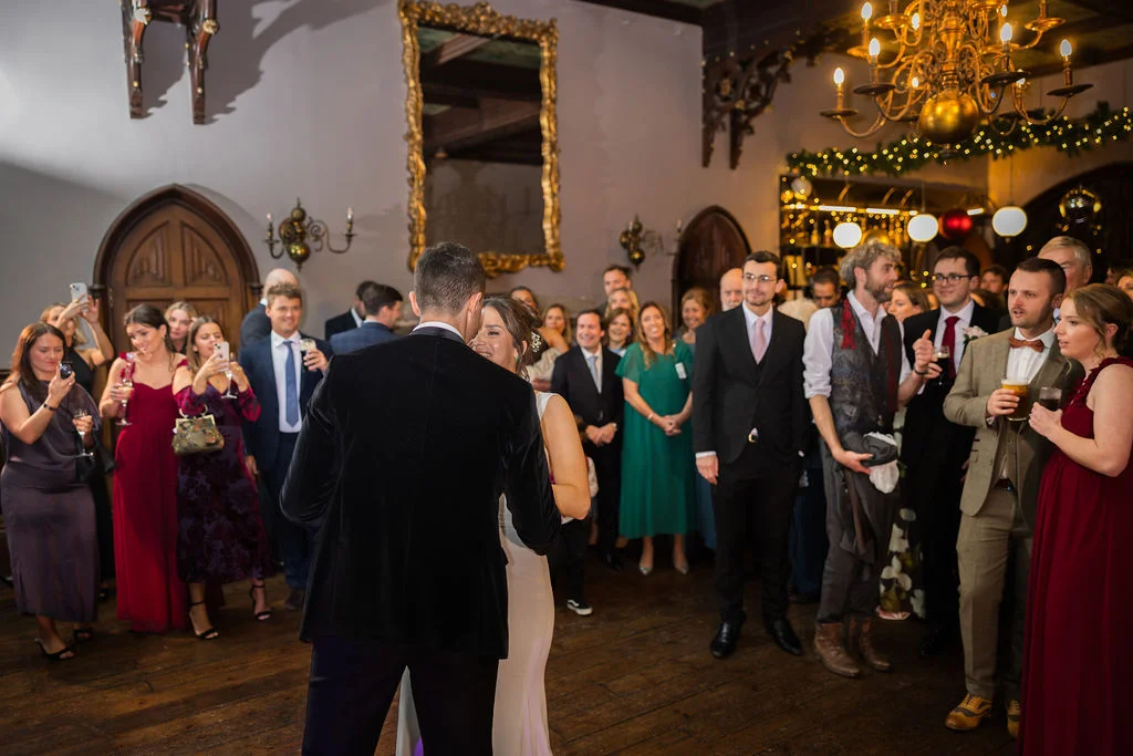 The newlywed couple shares their first dance on a polished wooden floor in a grand manor house ballroom, surrounded by admiring wedding guests.