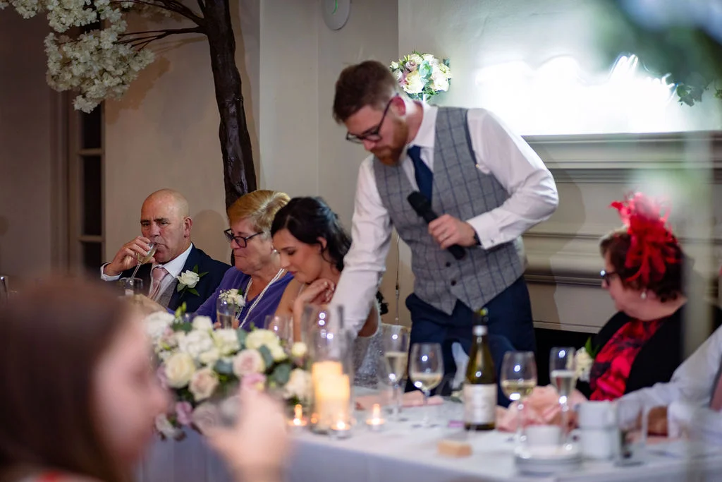 A groom delivers a speech holding a microphone at the top table during a wedding reception, surrounded by family members and floral arrangements.