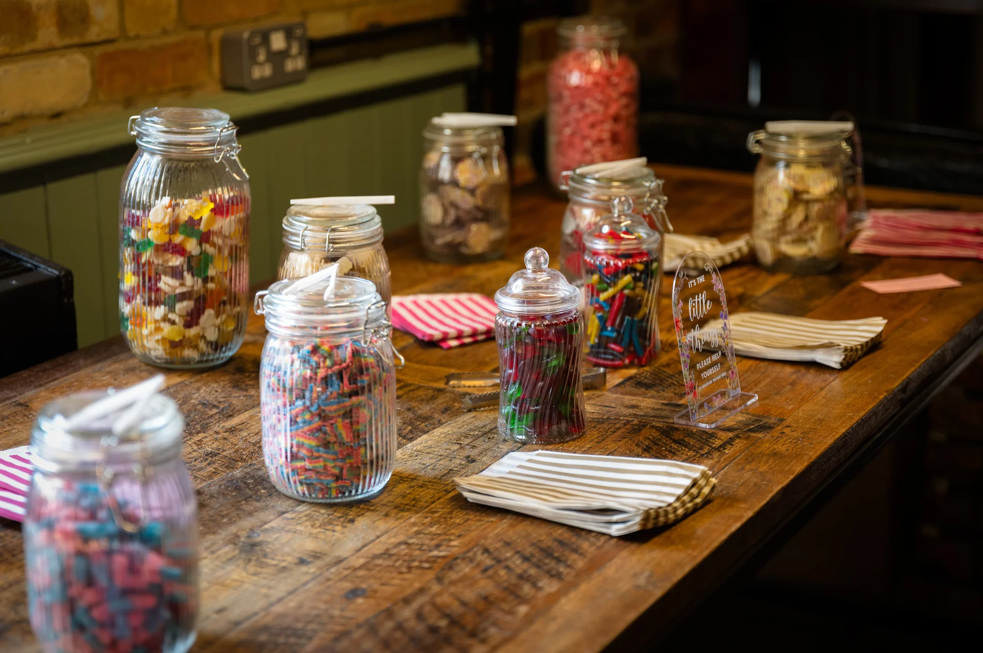 A rustic wooden table is arranged with multiple glass sweet jars filled with colourful pick and mix sweets, accompanied by stacks of napkins and a small acrylic sign at a wedding reception.