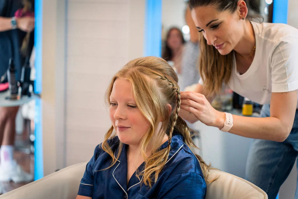 A junior bridesmaid with soft waves and braided hair details gets her hair styled while wearing a blue satin top during morning preparations.