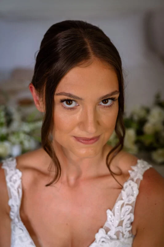 Close-up portrait of a radiant bride with soft brown hair in an updo and face-framing tendrils, wearing a lace wedding dress with a sweetheart neckline.