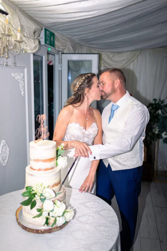 The newlywed couple share a tender kiss while cutting their semi-naked three-tier wedding cake decorated with white floral accents inside a draped marquee.