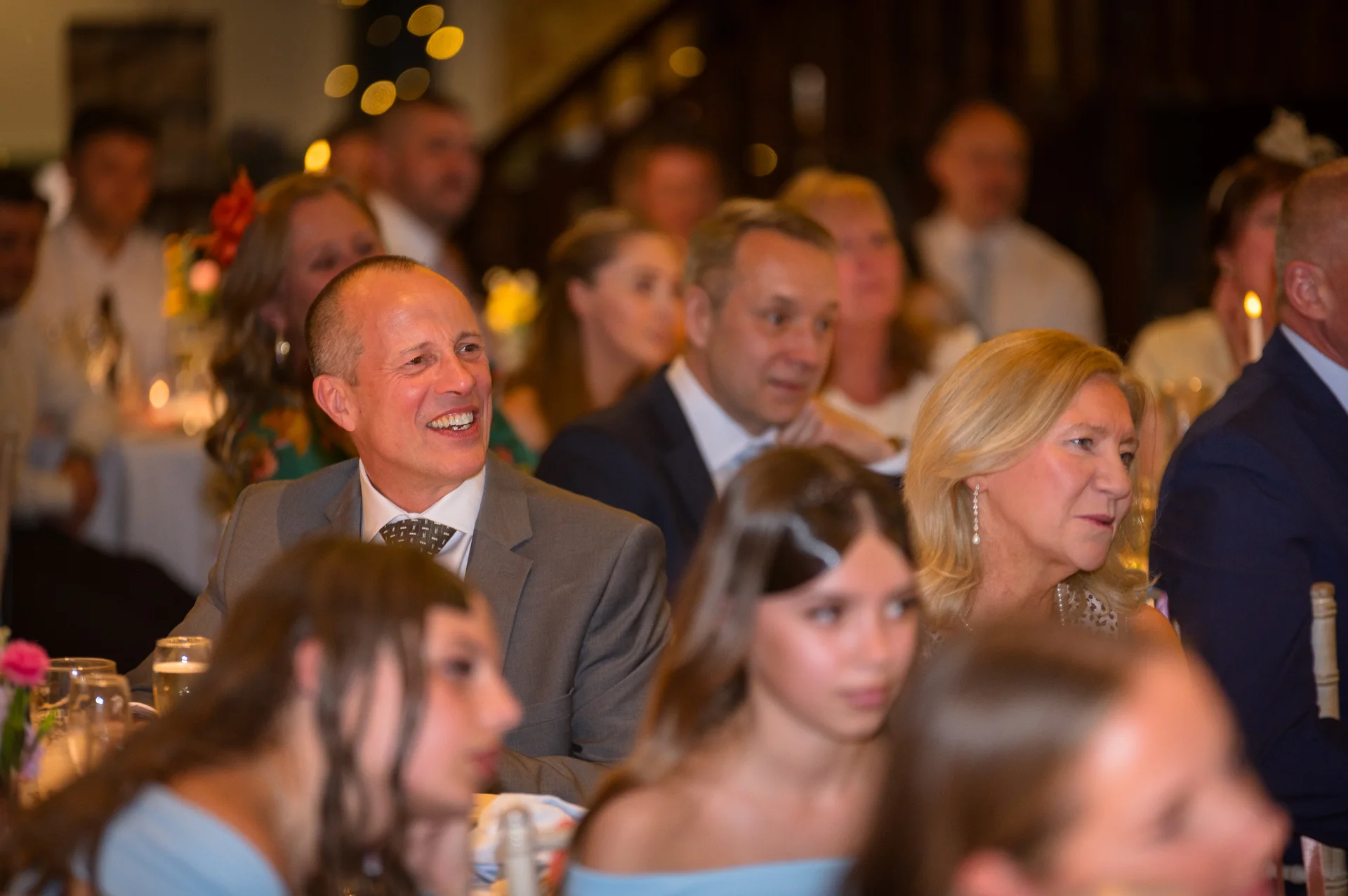 A wedding guest in a grey suit laughs joyfully during the reception speeches while seated amongst an audience of family and friends.