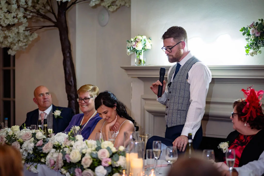 The groom stands at the top table delivering a speech, holding a microphone while the bride smiles bashfully beside him amidst floral table arrangements and family members.