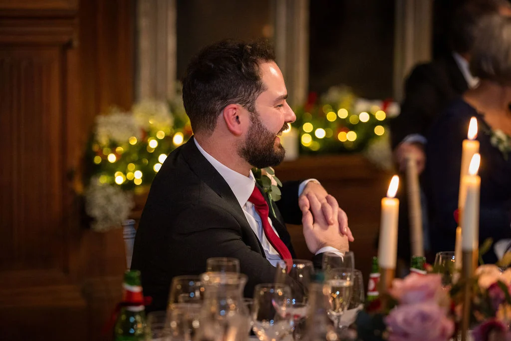 A smiling groom in a black suit and red tie sits at a candlelit wedding reception table while listening to speeches.