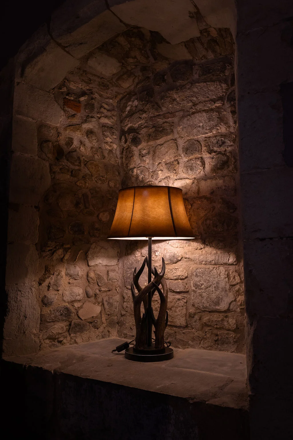 A warm, glowing table lamp with an antler-style base rests inside a rustic stone alcove, casting soft amber light against the textured wall.