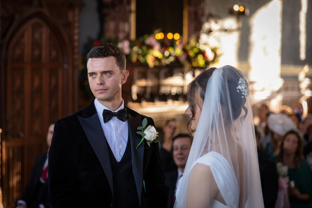 A groom in a black velvet tuxedo looks away attentively while standing next to his bride during their church ceremony.