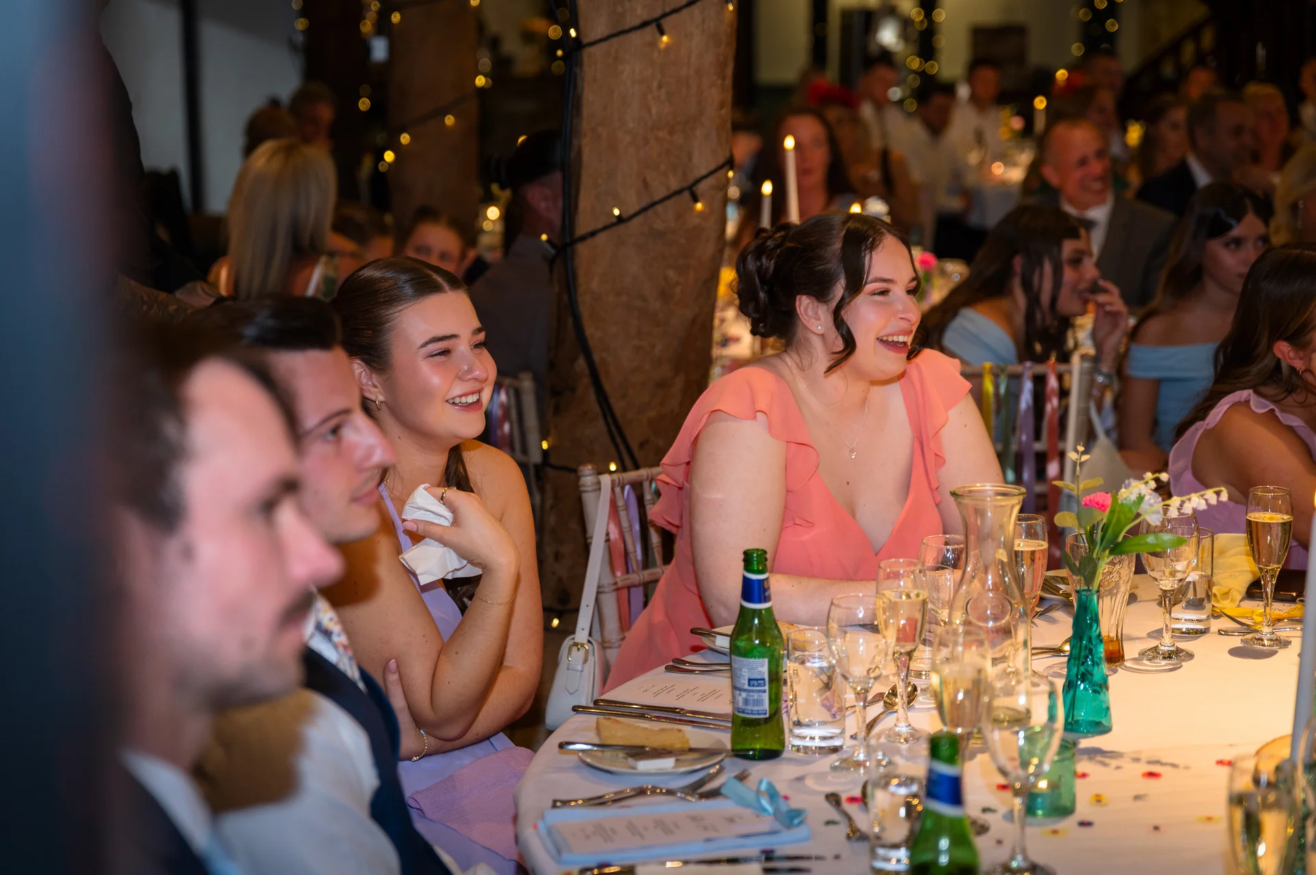 Wedding guests seated at a long banquet table inside a rustic barn are laughing happily during speeches.