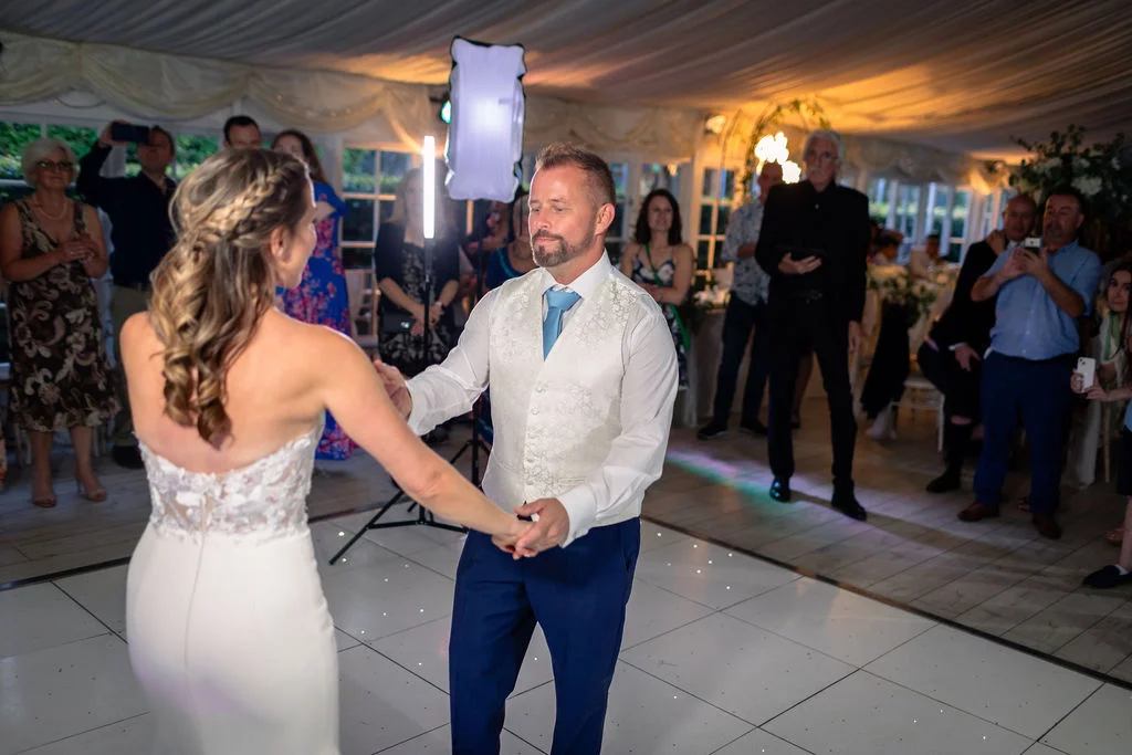 Newlyweds share their first dance on a glowing dance floor inside a marquee reception while wedding guests watch from the sidelines.