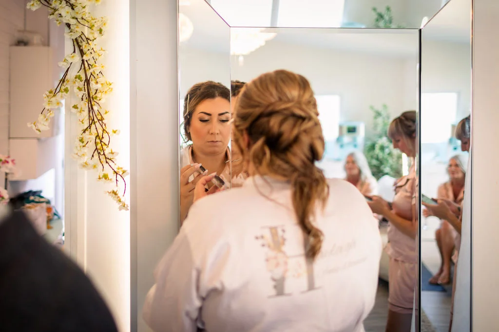 A bride stands in front of a multi-panel mirror during morning preparations while wearing a personalised getting-ready robe, with her bridesmaids visible in the reflection.