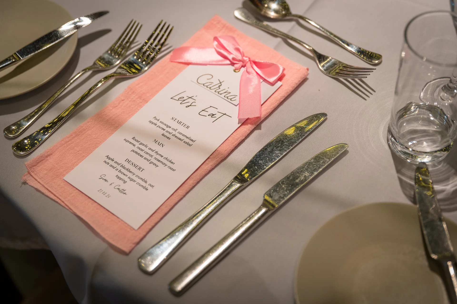 A close-up of a wedding place setting featuring a personalised menu card tied with a pink ribbon on a peach napkin and crisp tableware.