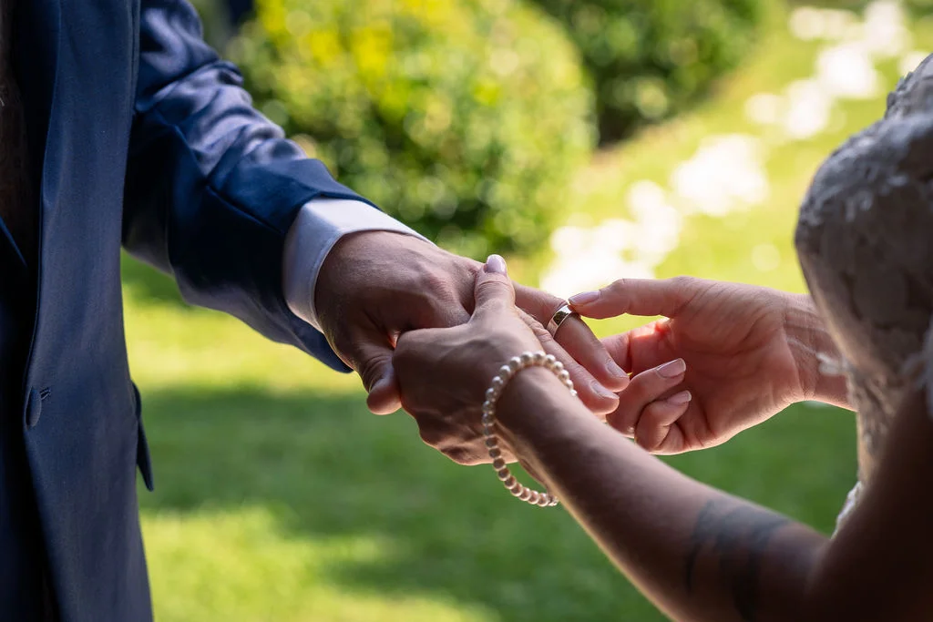 Close-up of a bride placing a wedding ring onto the groom's finger during an outdoor ceremony, with lush greenery in the blurred background.