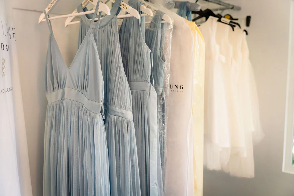 A selection of pleated dusty blue bridesmaid dresses and white garments hang on matching hangers during morning bridal preparations.