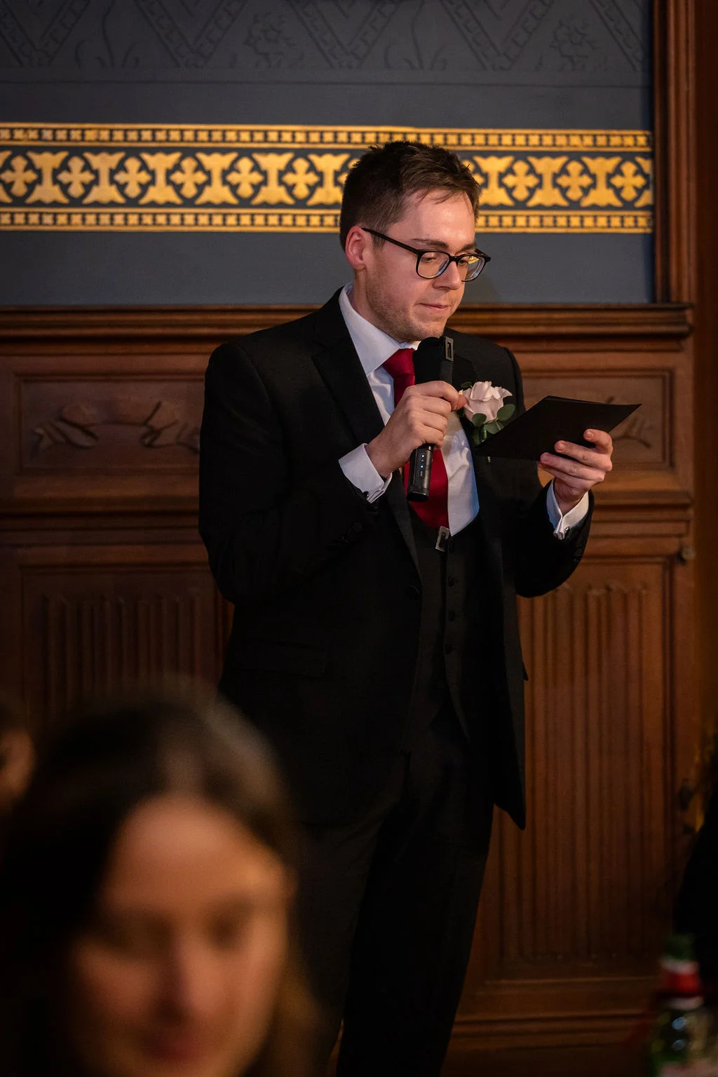 A groom in a black three-piece suit with a red tie delivers his speech, holding notes and a microphone in a wood-panelled room.