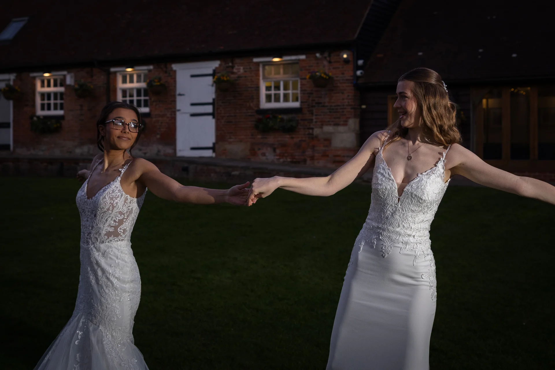 Two smiling brides in elegant white lace wedding gowns hold hands and playfully spin together on the lawn outside a rustic brick barn at twilight.