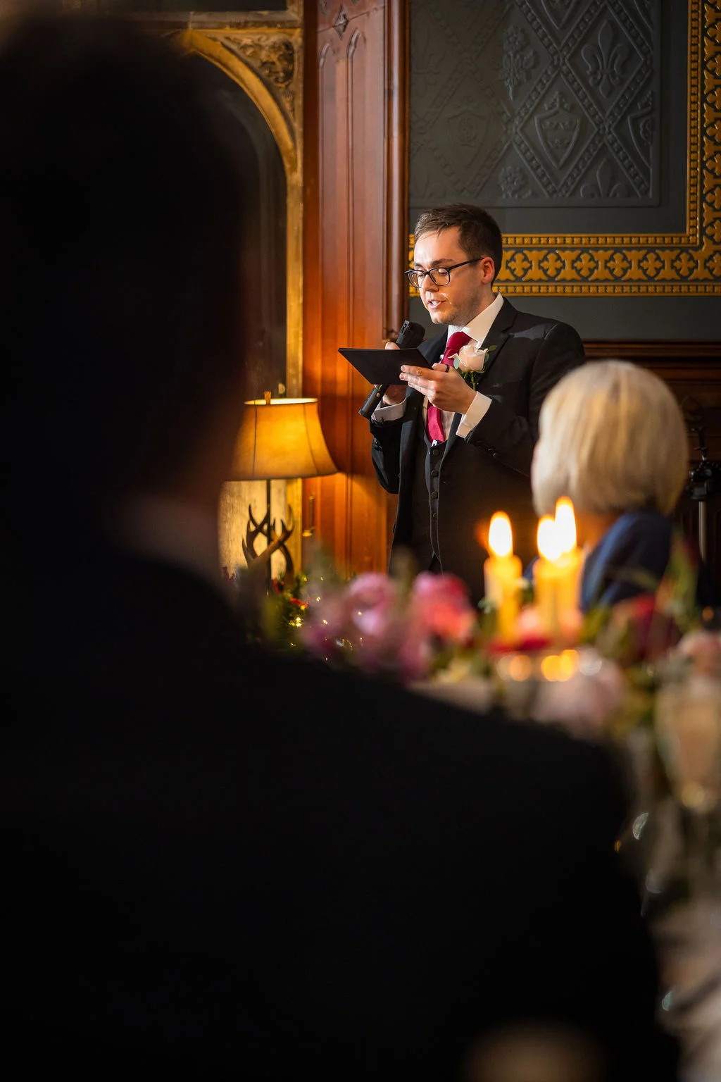 A best man delivers an emotional speech holding notes during a candlelit wedding reception in a wood-panelled country house.