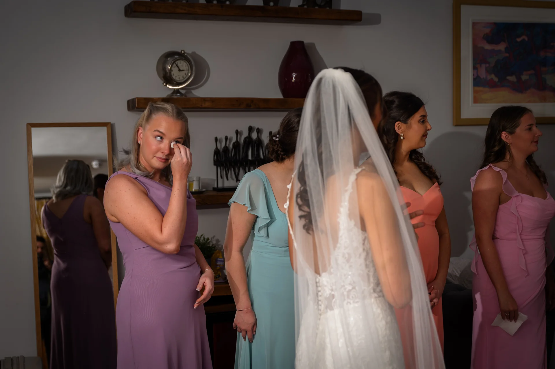 An emotional bridesmaid wipes away a tear while watching her friend in a lace wedding dress and veil during the morning preparations.