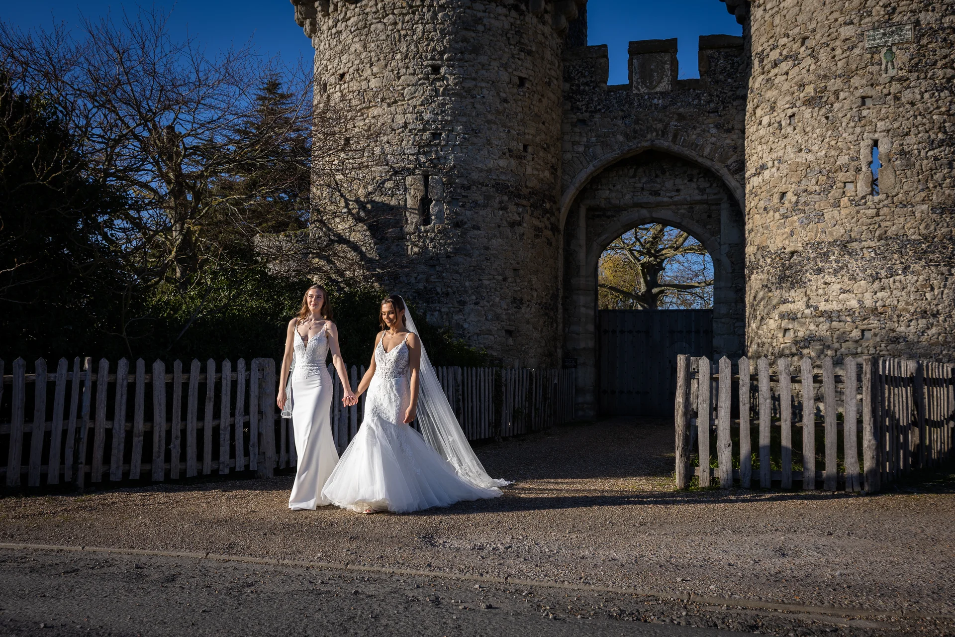 Two brides walk hand in hand outside a historic stone castle on a bright, sunny wedding day.