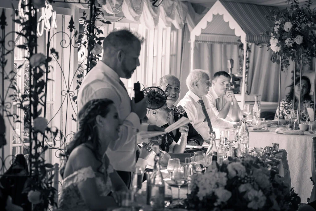 A groom delivers a speech at the top table during a marquee wedding reception while guests listen and laugh.