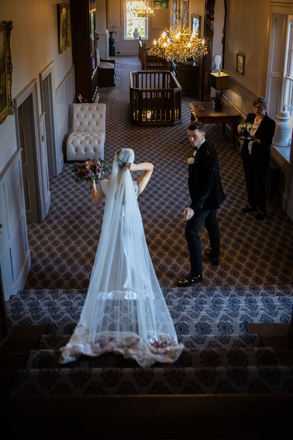 A bride standing on a grand hotel staircase adjusts her chapel-length veil while the groom looks back and a staff member holds a tray of drinks nearby.