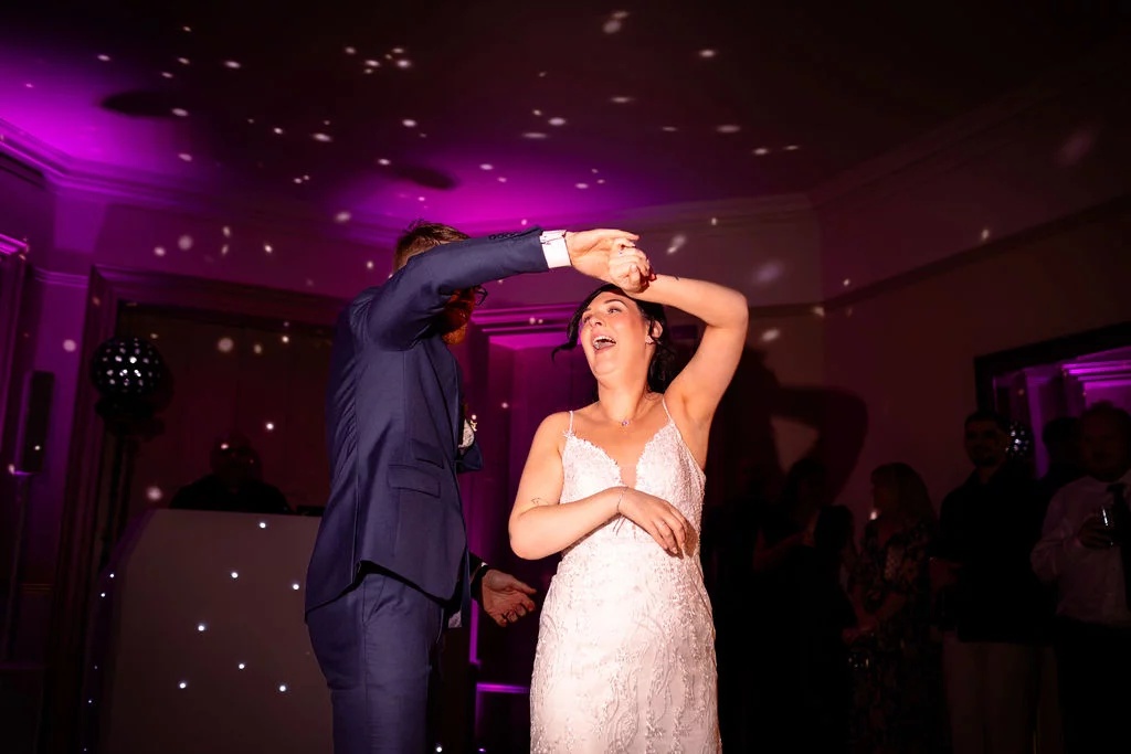 The bride and groom share a joyful spin on the dance floor beneath pink lighting and sparkling disco lights, surrounded by cheering guests.