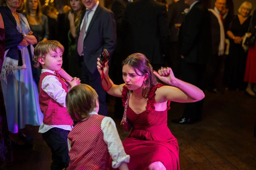 A guest in a red dress dances playfully with children on the wooden dance floor during an evening wedding reception.