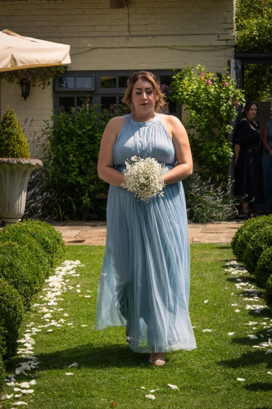 A bridesmaid walks down a garden aisle lined with white petals, wearing a dusty blue halter-neck chiffon dress and holding a bouquet of baby's breath.