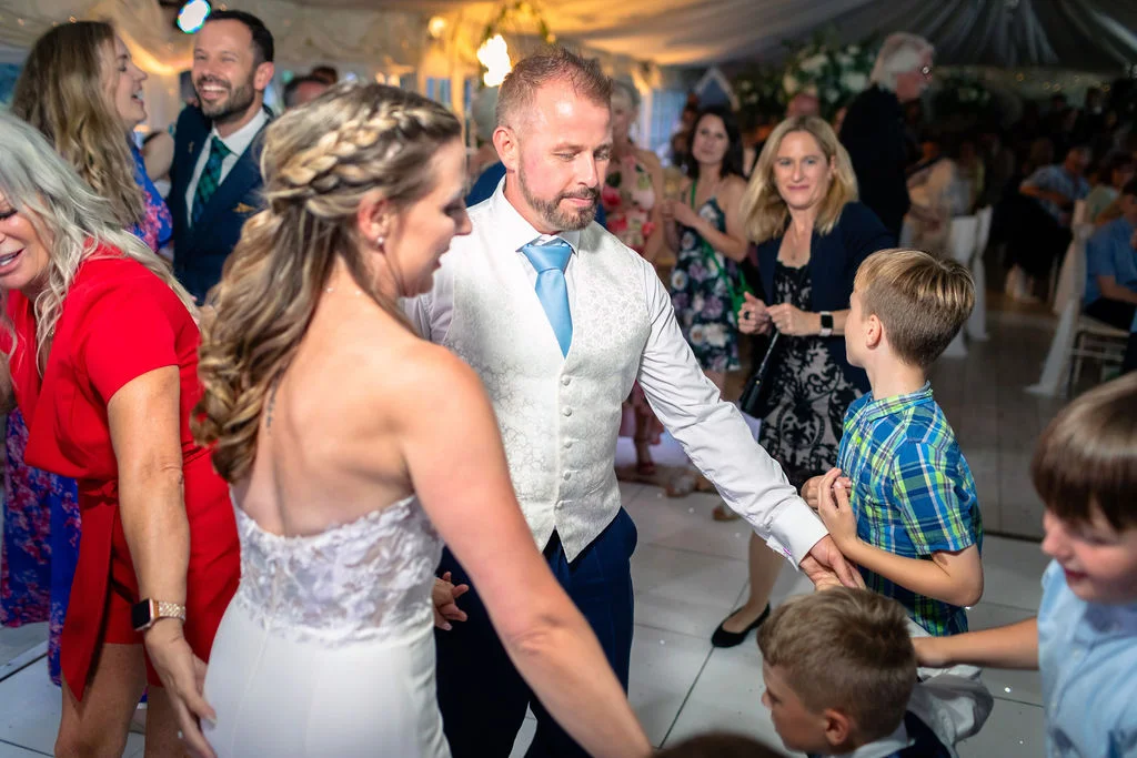 A newlywed couple joyfully dances in a crowded marquee reception surrounded by smiling guests and children.