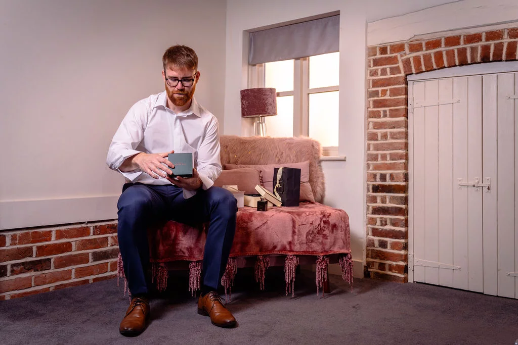 A groom in a white shirt and trousers sits on a pink velvet bench while opening a gift box during wedding morning preparations.