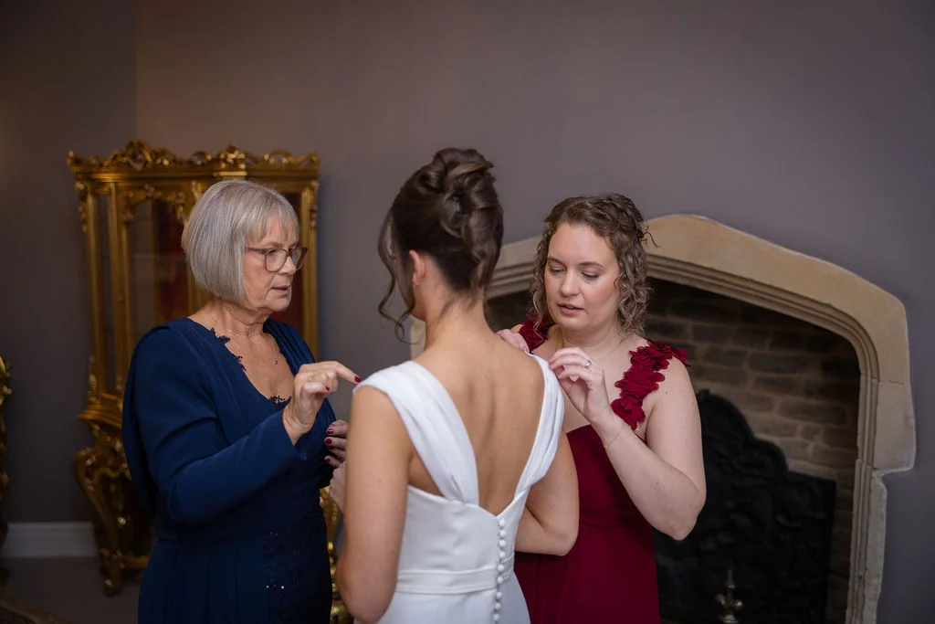 The mother of the bride and a bridesmaid assist the bride with the final touches to her wedding dress during morning preparations.