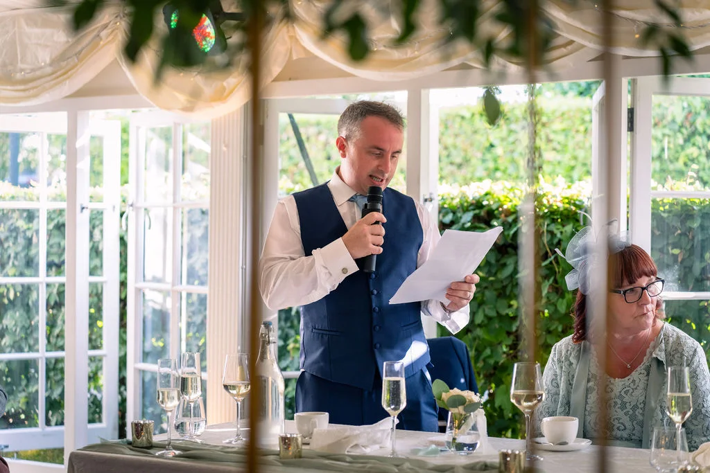 A groom delivers his speech during the wedding reception inside a brightly lit marquee decorated with white ceiling draping and rustic table settings.