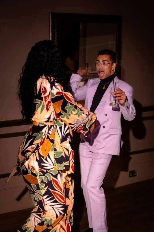 A guest in a vibrant patterned outfit dances joyfully with a man in a stylish lilac double-breasted suit holding a glass on the dance floor.