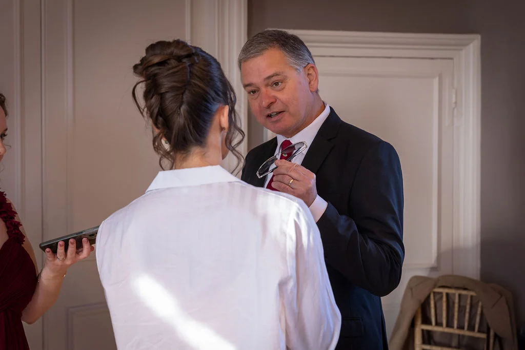 A father in a black suit holds glasses while sharing a tender moment with his daughter who is seen from behind with an updo hairstyle during morning wedding preparations.