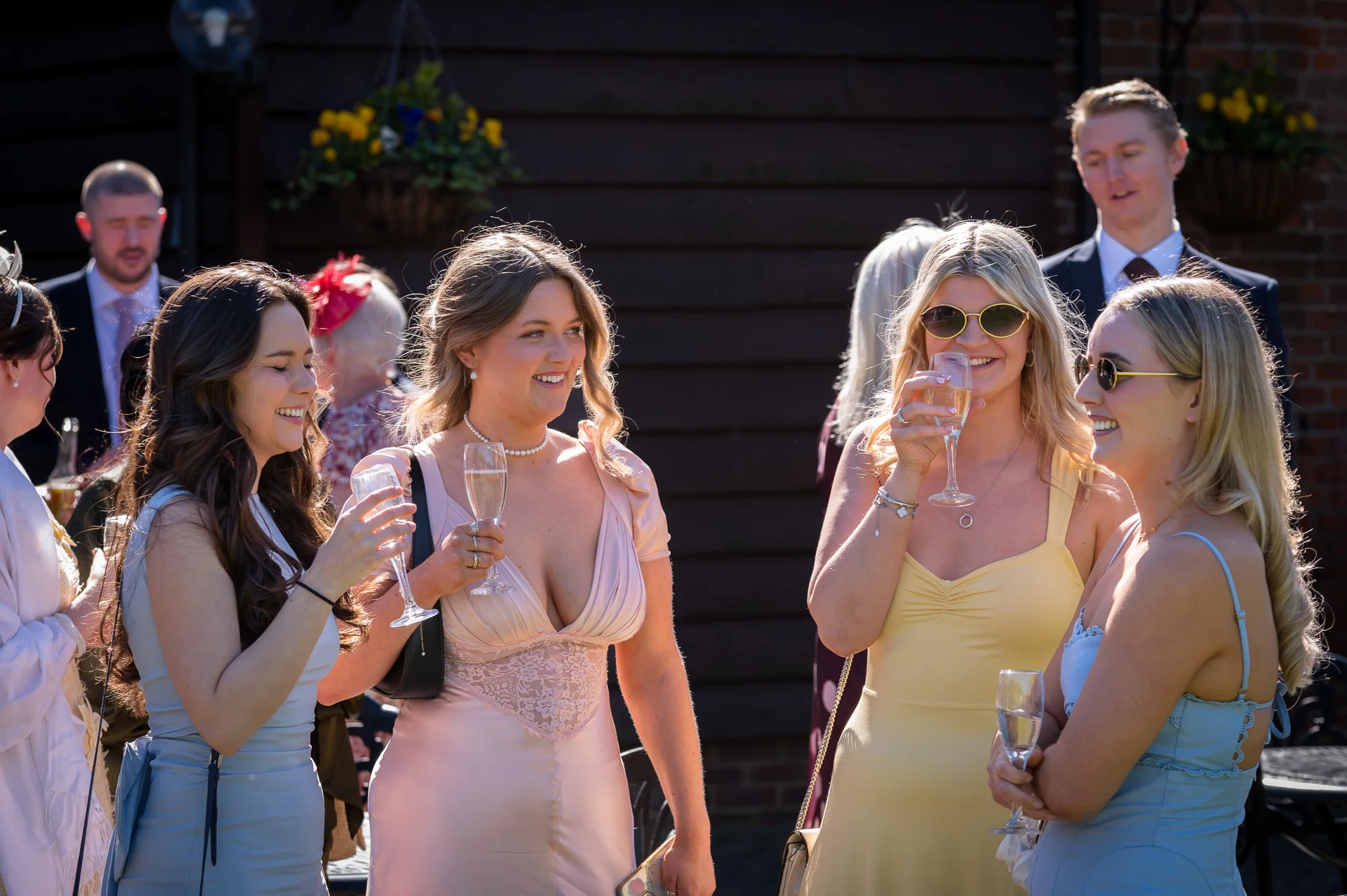 A group of wedding guests mingle outdoors in the sunshine, holding glasses of champagne and laughing together during the drinks reception.