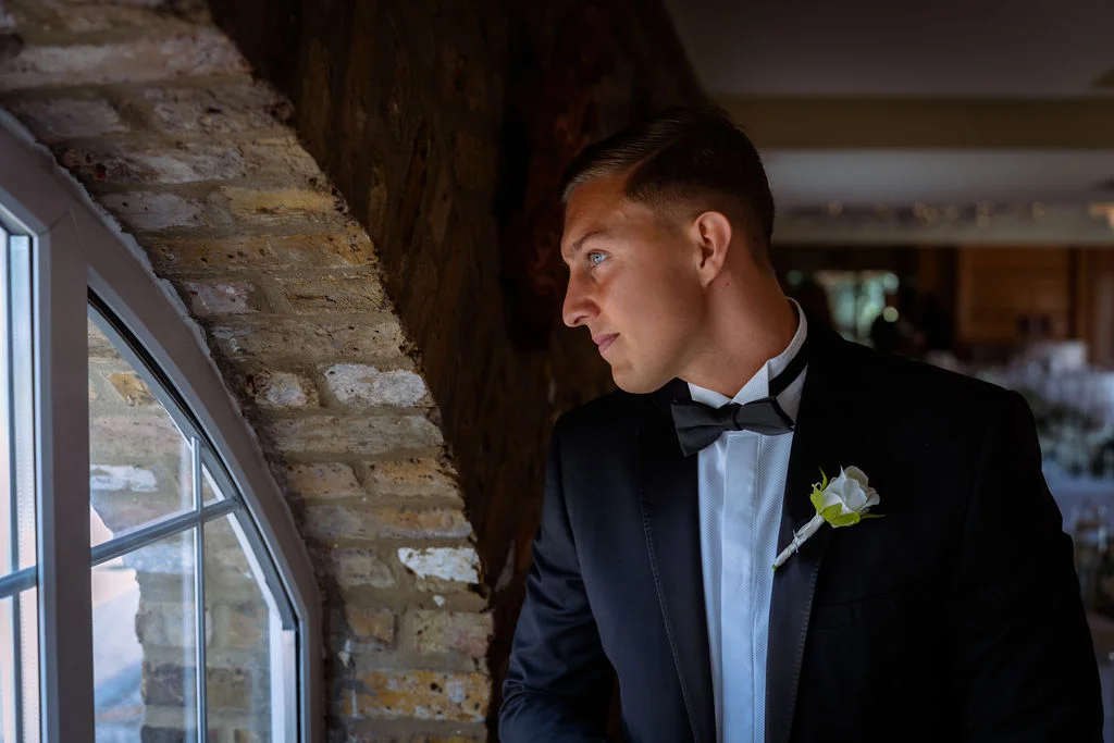A handsome groom in a classic black tuxedo and bow tie gazes thoughtfully out of an arched window framed by exposed brick.