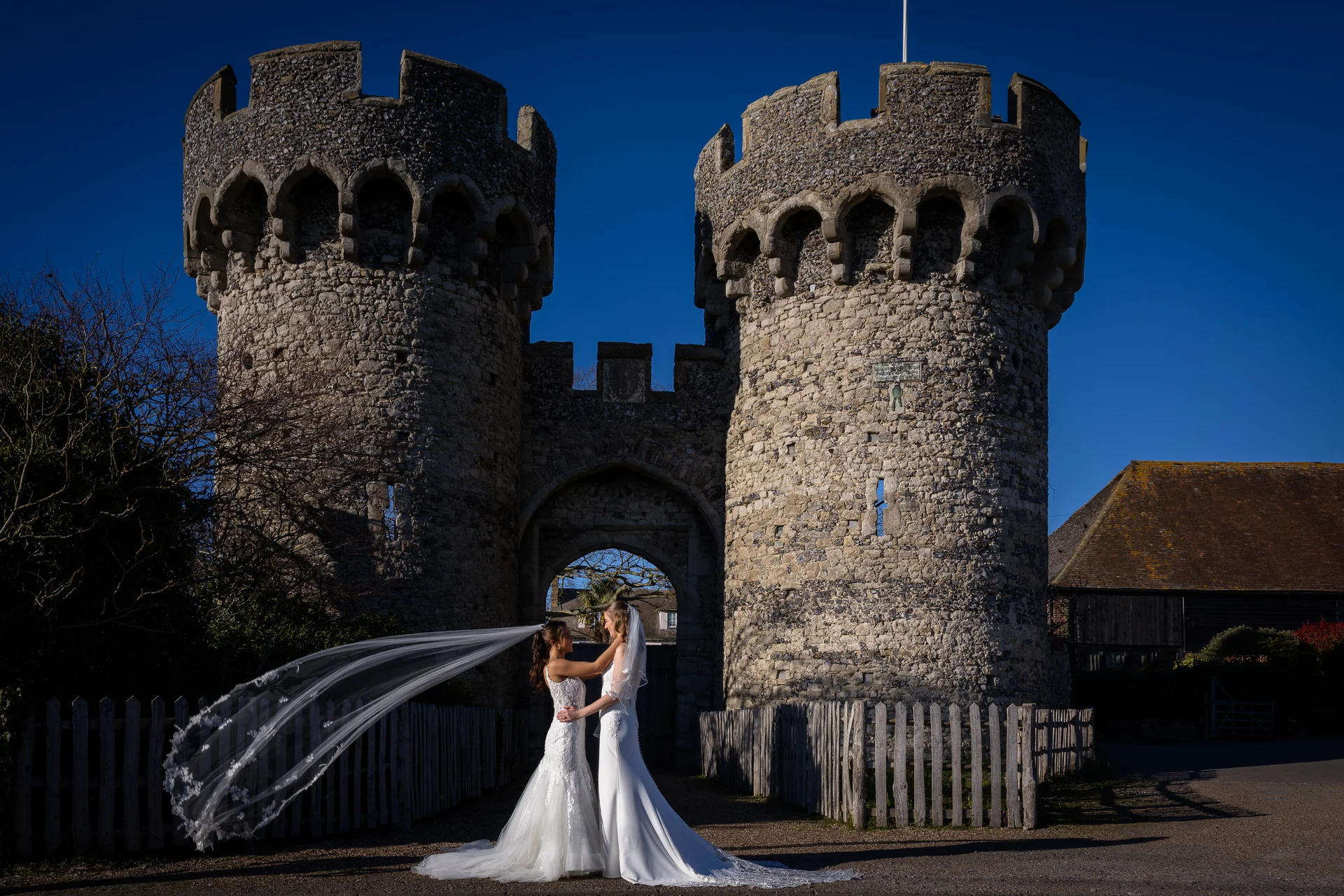 Two brides in white wedding gowns embrace in front of historic stone castle towers while a long veil billows dramatically in the wind under a clear blue sky.