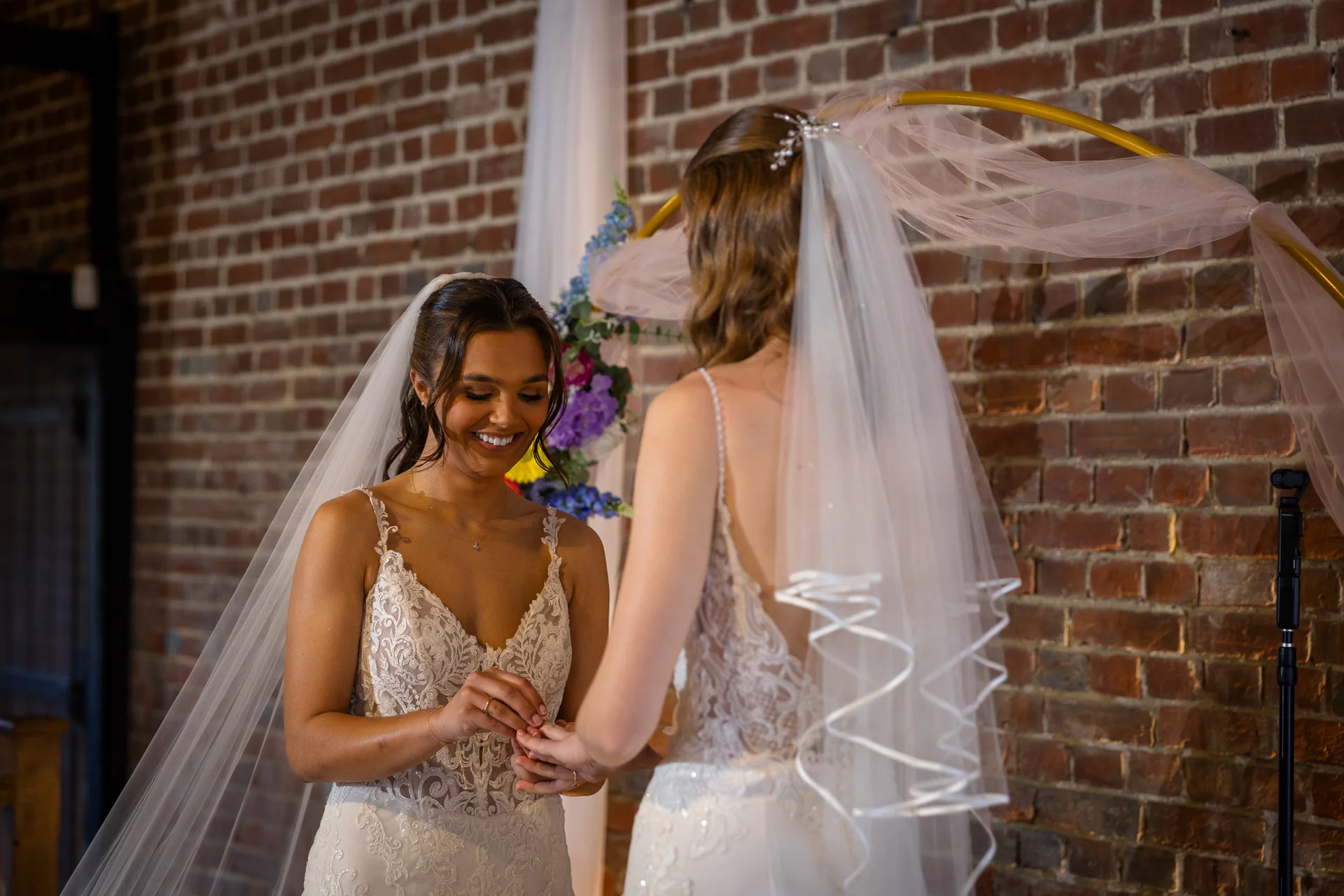 Two brides smiling joyfully as they exchange rings during their wedding ceremony in front of an exposed brick wall and a floral arch.