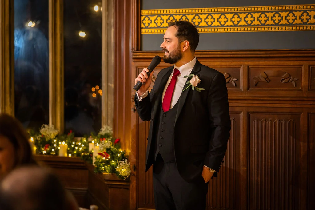 A groom in a three-piece black suit and red tie delivers a speech into a microphone inside a wood-panelled room.
