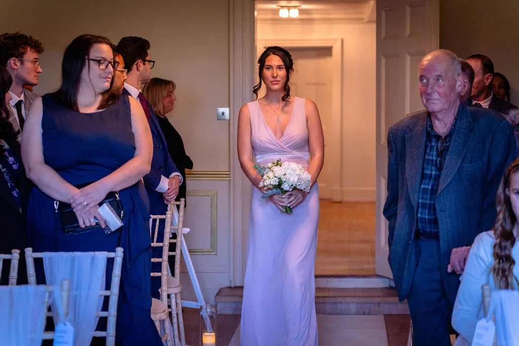 A bridesmaid wearing a pink v-neck dress and holding a white posy bouquet walks down the aisle during a wedding ceremony.