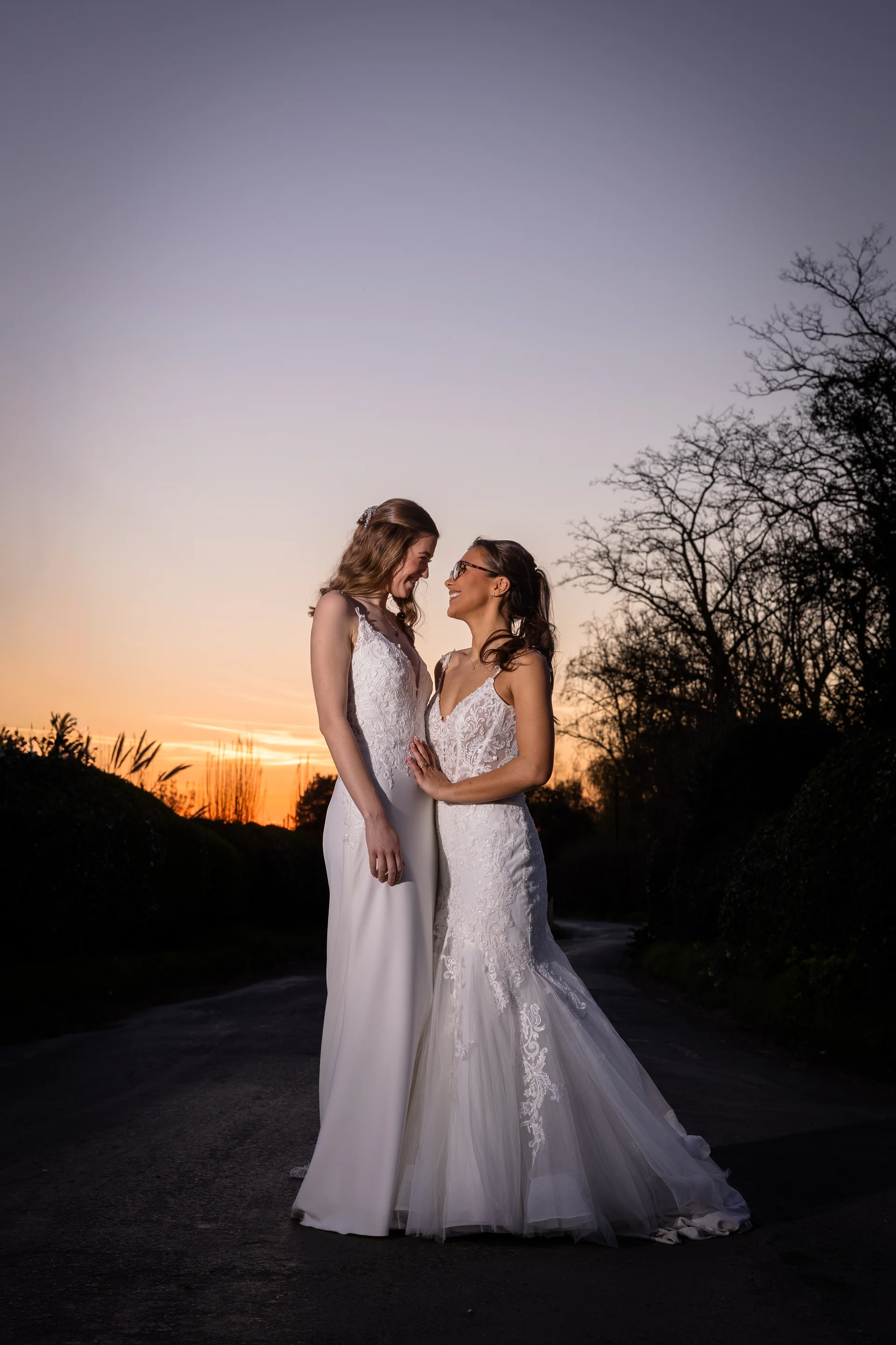 Two brides in white lace wedding dresses share a tender moment outside at dusk against a glowing sunset sky.