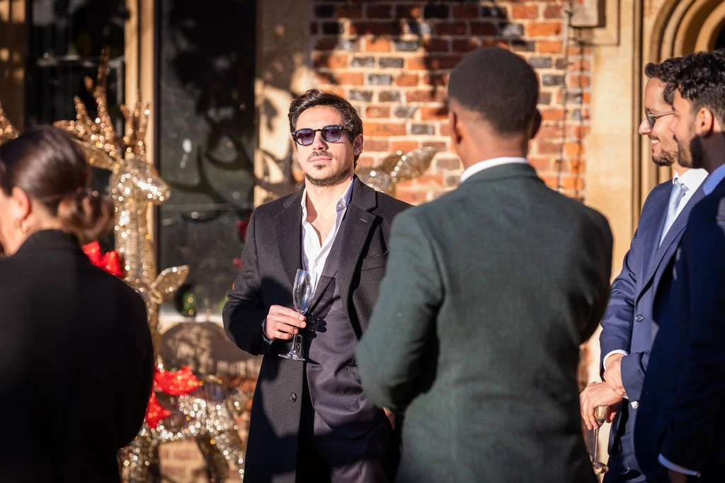 A group of smartly dressed wedding guests mingle outdoors in the warm golden hour light, holding champagne glasses and wearing sunglasses.