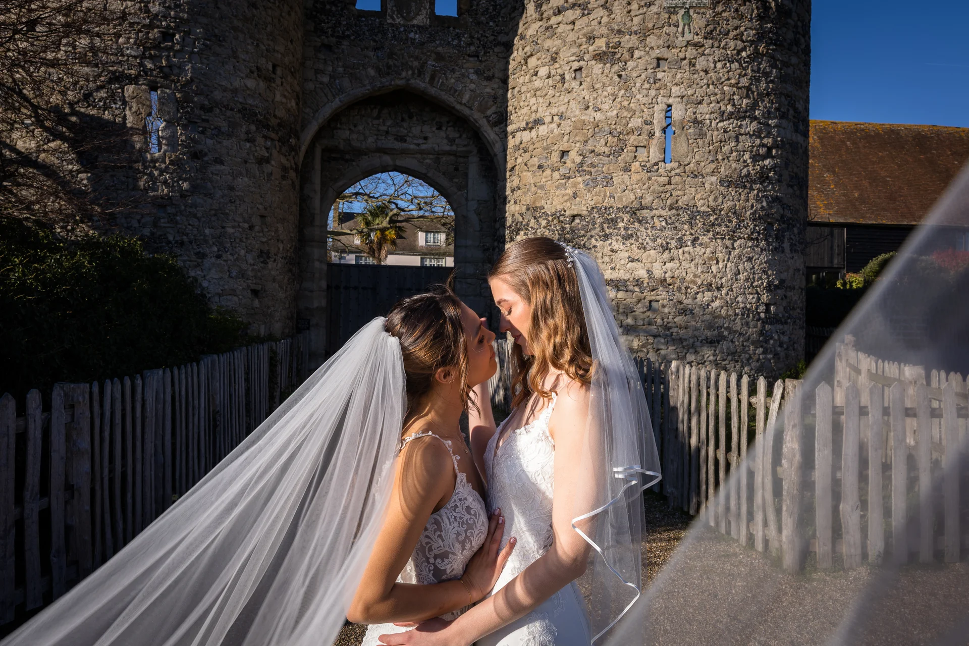 Two brides in lace wedding dresses and veils share a tender, intimate moment outside a historic stone castle.