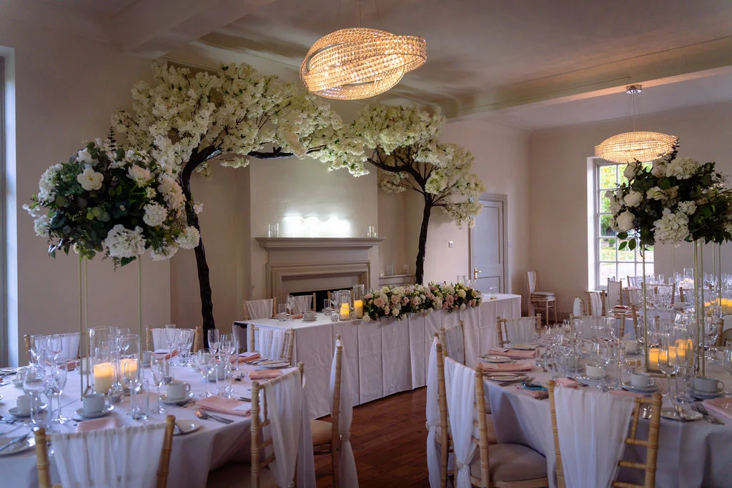 An elegantly decorated wedding reception room features round and long banquet tables adorned with white linens, tall floral centrepieces of roses and hydrangeas, and striking blossom tree installations framing the top table.