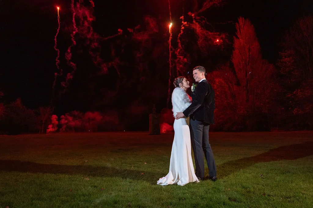 A newlywed couple embraces and laughs on a dark lawn under a dramatic sky illuminated by vibrant red fireworks.